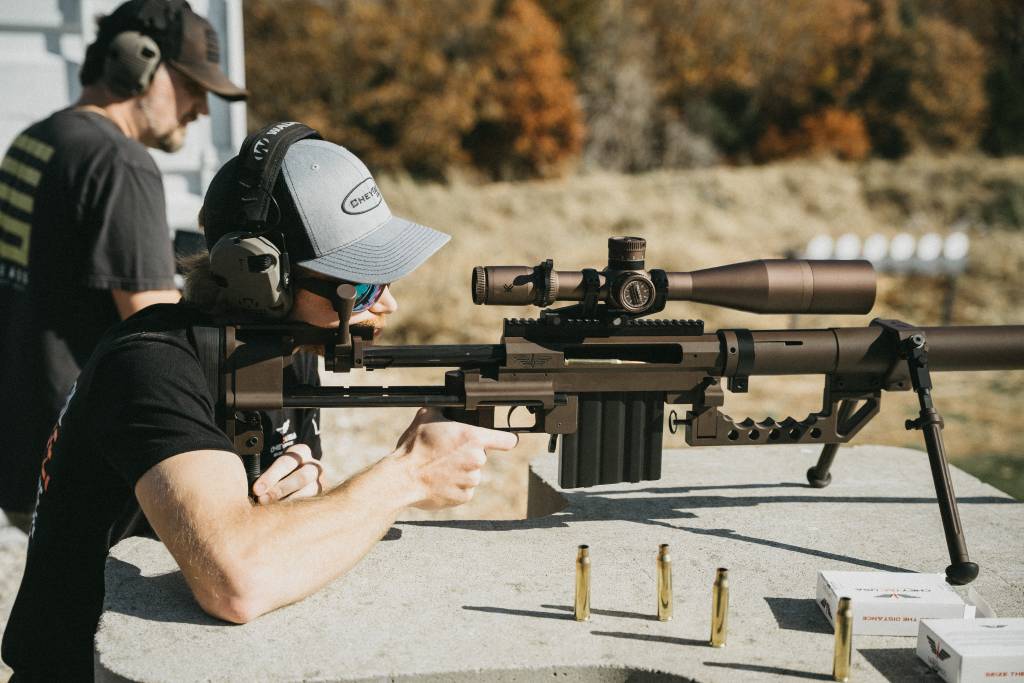 A man looking down the scope of an M200 intervention rifle with .408 casings next to him. 